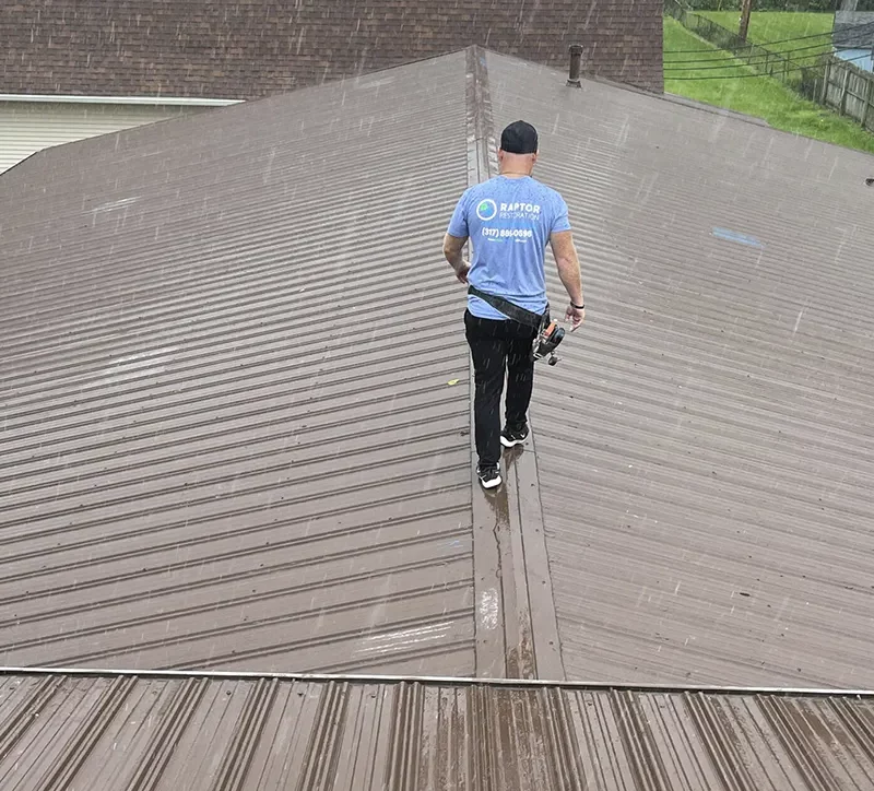 Raptor Roofing team member inspecting a ribbed metal roof during rainfall